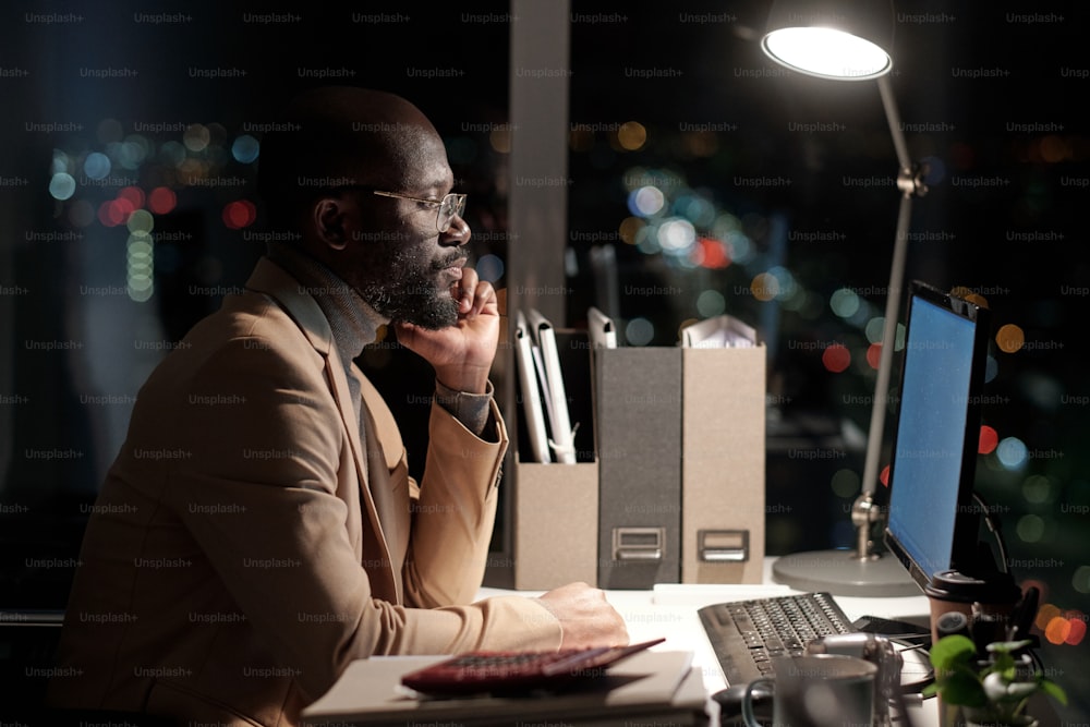 Side view of serious businessman of African ethnicity looking at computer screen by his workplace in office