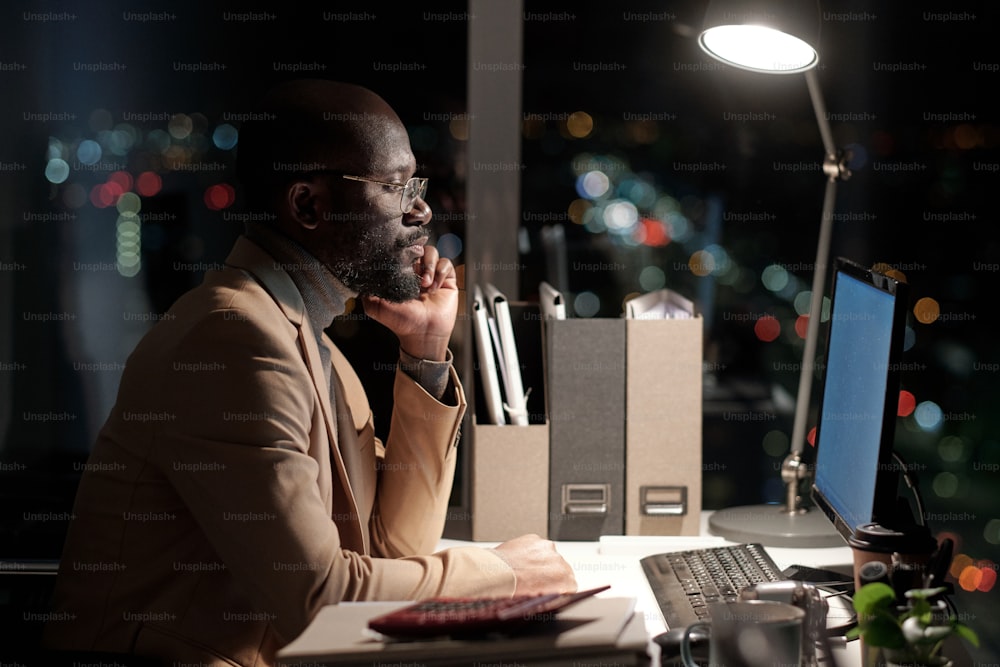Vista lateral de empresário sério de etnia africana olhando para a tela do computador por seu local de trabalho no escritório
