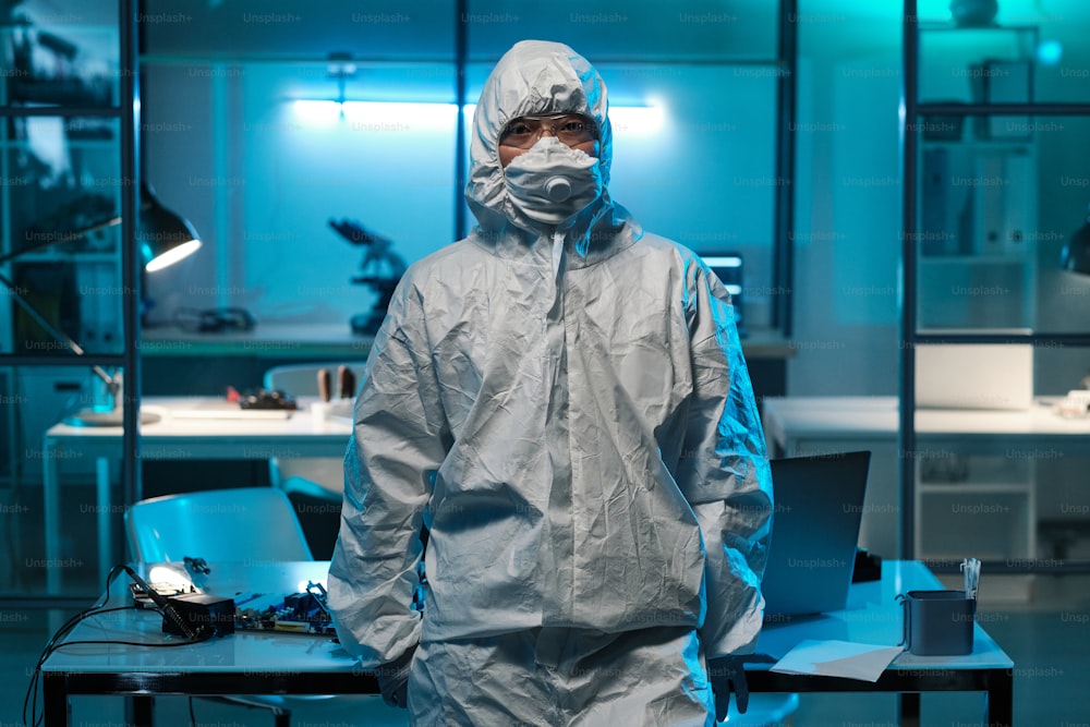 Jeune technicien ou scientifique contemporain en tenue de travail de protection debout contre un bureau avec un ordinateur portable et du matériel