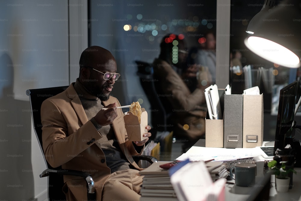 Hungry young businessman with chopsticks and box of chinese wok sitting by workplace in front of computer monitor