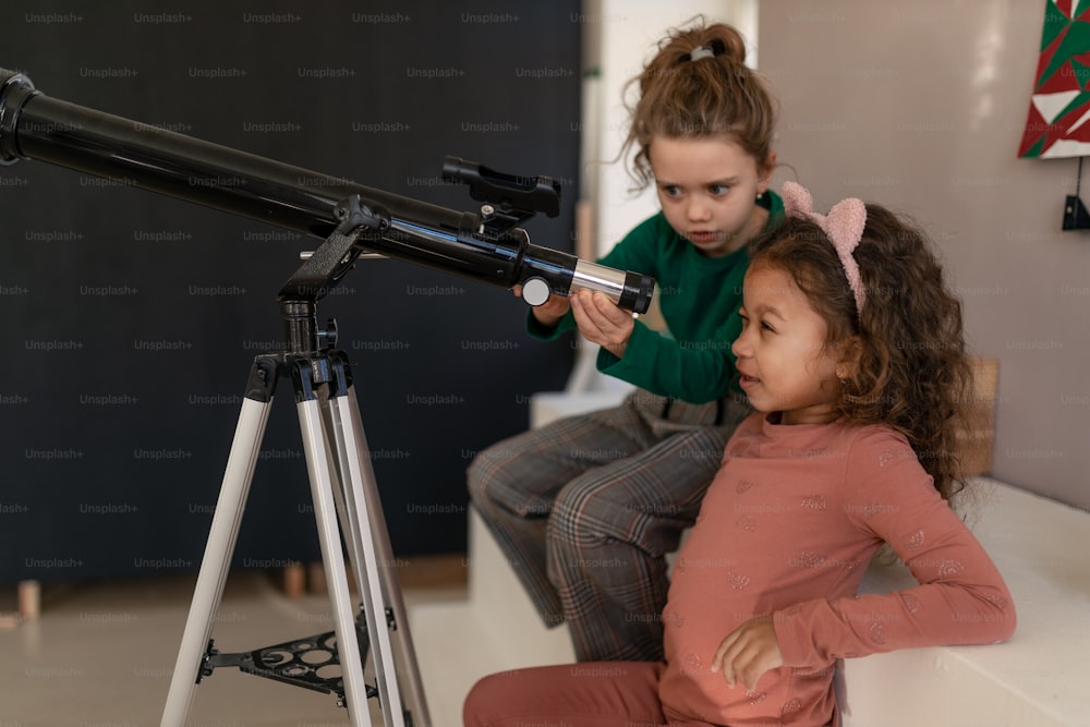 Duas meninas olhando através de um telescópio dentro de casa.