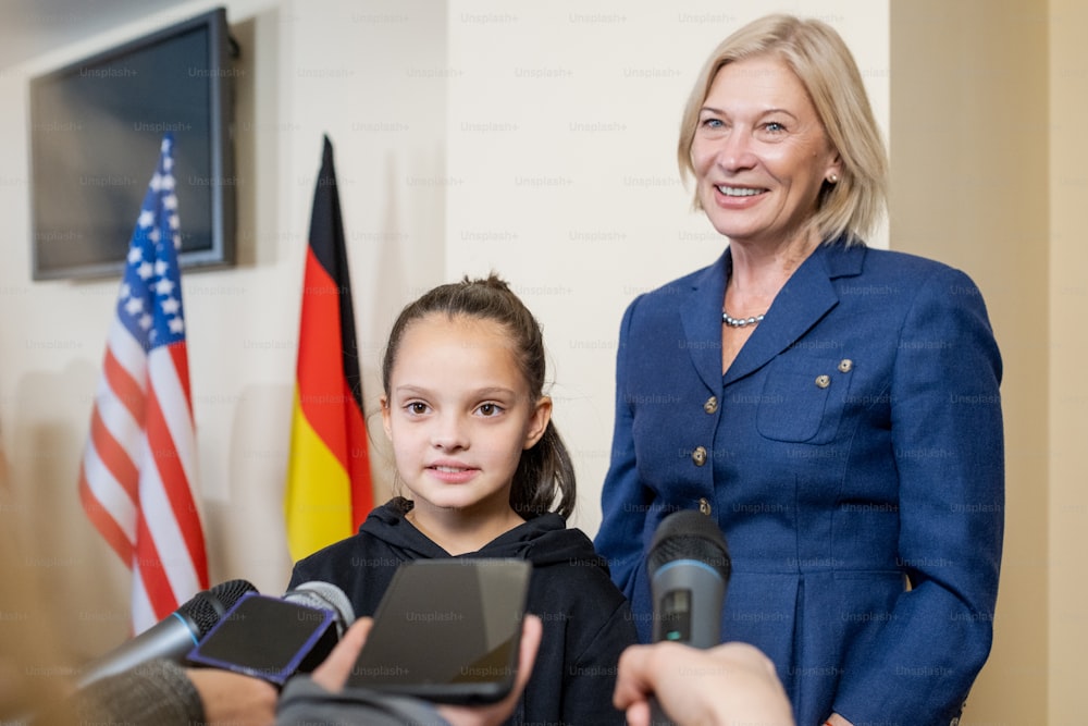 Schoolgirl and mature blond female answering questions of journalists during press-conference in large auditorium