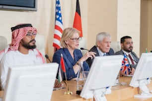 금발의 성숙한 여성 대표가 여러 외국 동료들 사이에 테이블에 앉아 청중을 위해 연설을 하고 있다