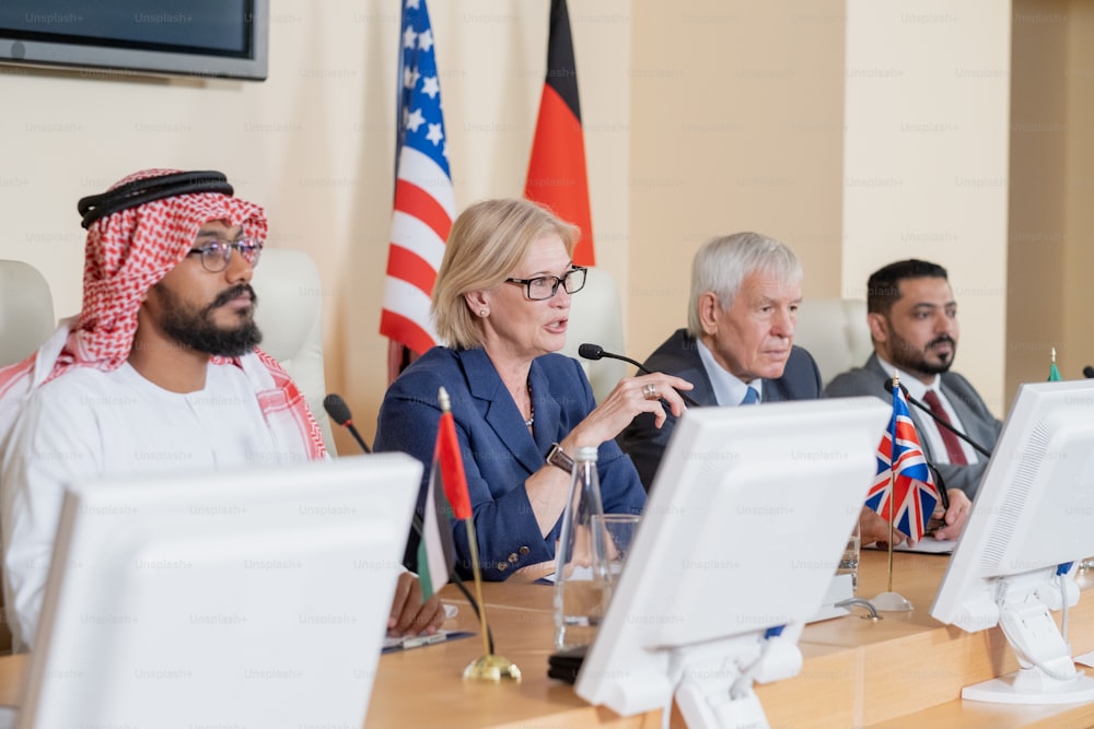 Blond mature female delegate making speech for audience while sitting by table between several foreign colleagues