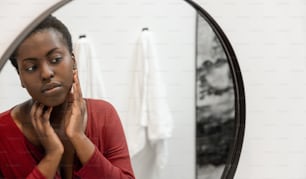 Reflejo en el espejo de una mujer africana negra tocando e inspeccionando su rostro. Cuidado personal y cuidado de la piel