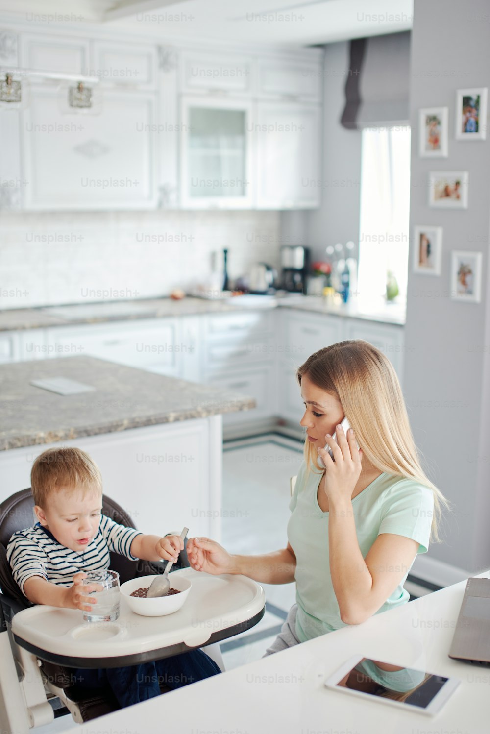 Porträt einer mittelalten kaukasischen blonden Frau mit Kind, die am Telefon beschäftigt ist und das Baby mit Löffel füttert. Sie sind zu Hause und tragen Pyjamas