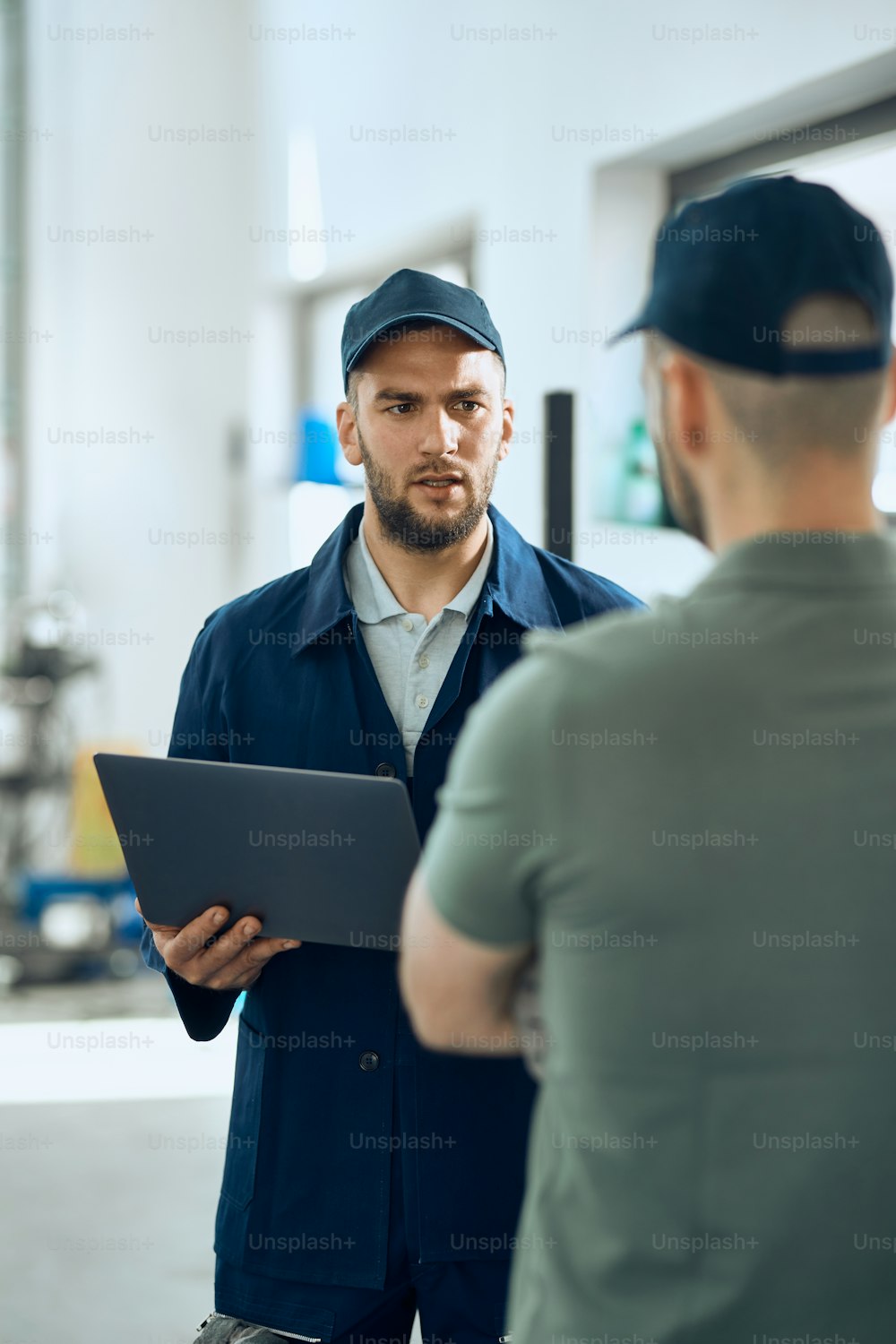 Young car mechanic giving instructions to his coworker while using laptop in auto repair shop.