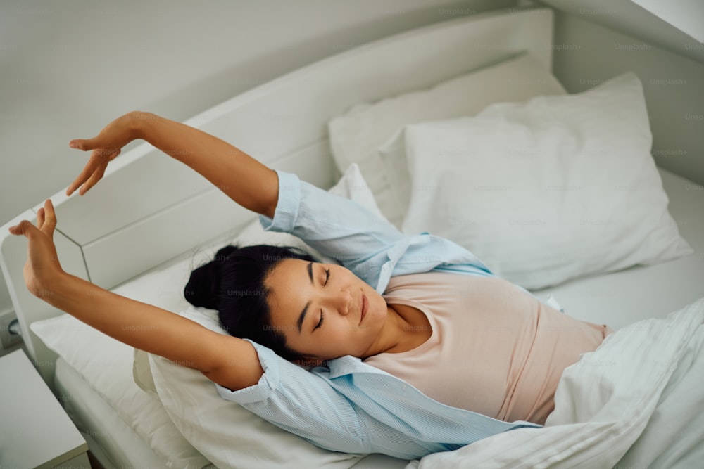 Jeune femme asiatique souriante se réveillant le matin et s’étirant sur un lit.