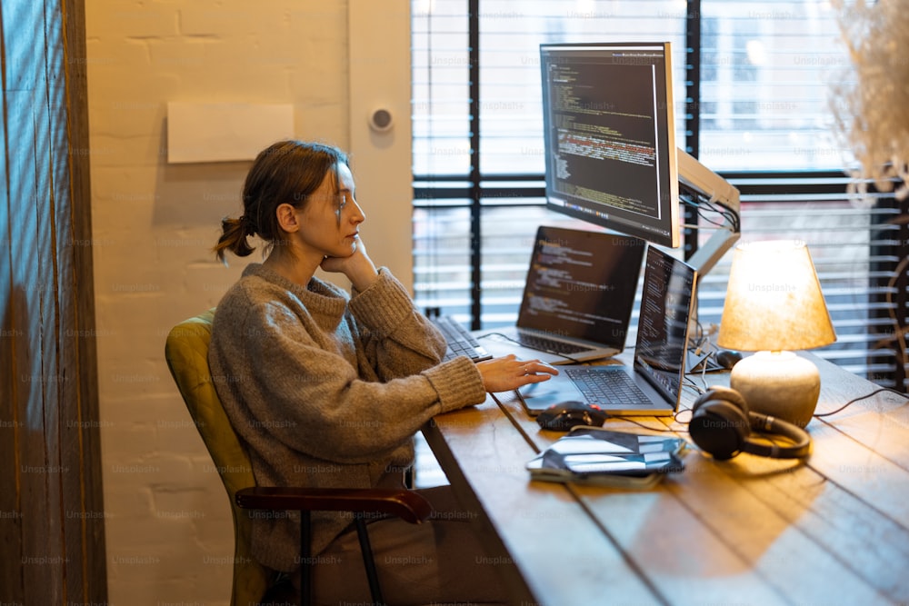 Young woman works on computers, sitting at workplace at cozy home office interior. Concept of freelance and remote work from home. Programmer writing code. Caucasian woman wearing domestic clothes.