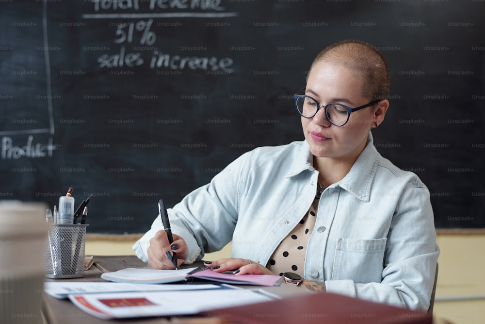 Young serious businesswoman in casualwear and eyeglasses making notes in notebook by workplace against blackboard