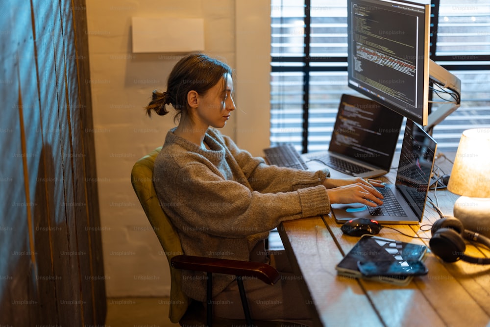 Young woman works on computers, sitting at workplace at cozy home office interior. Concept of freelance and remote work from home. Programmer writing code. Caucasian woman wearing domestic clothes.