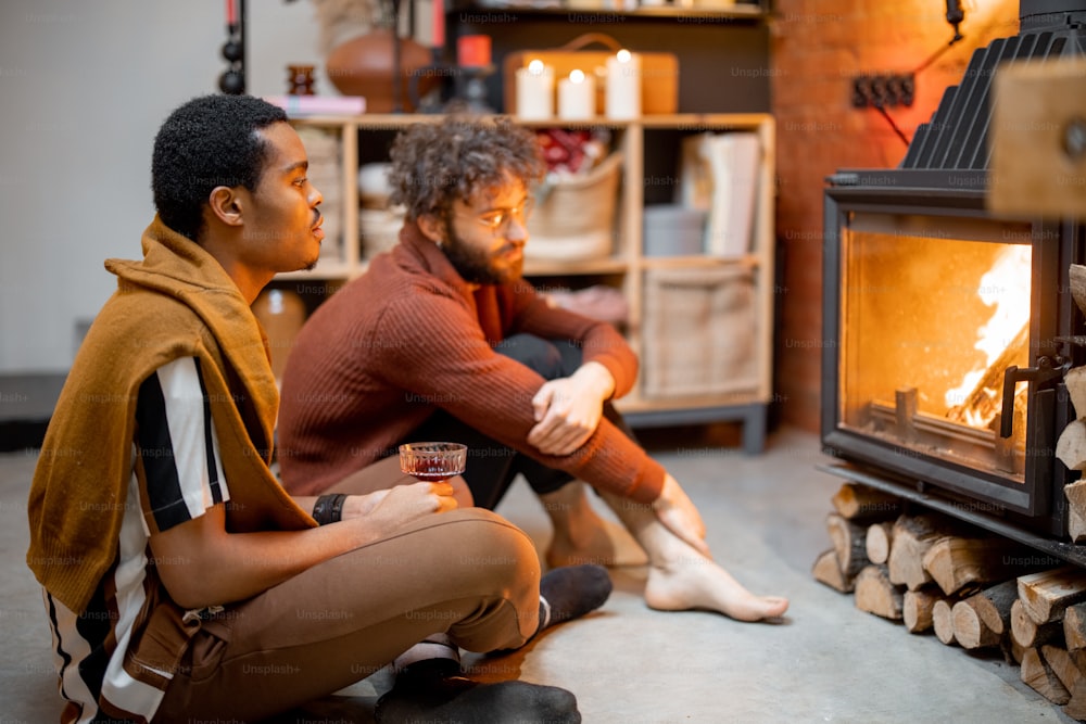 Two men sitting together by the burning fireplace at cozy home. Concept of homosexual relations and coziness on winter time. Idea of multinational gay families