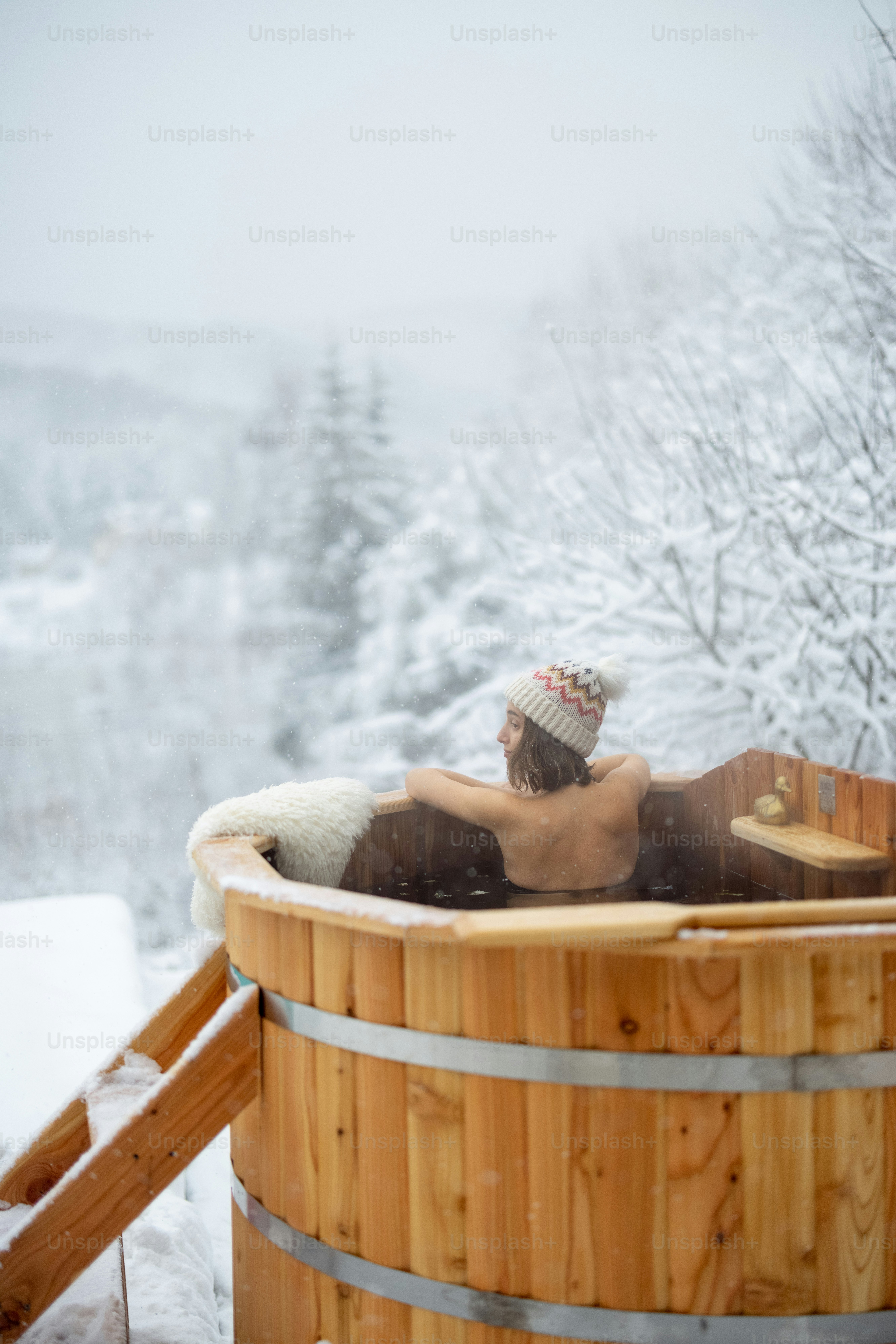 A person in a knit hat relaxing in a wooden hot tub outdoors in winter