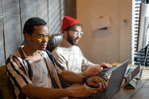 Two men with different nationality working on computers, sitting together at cozy home office. Concept of freelance and remote work. Stylish male hipsters programming together