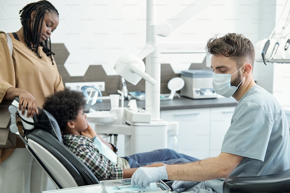 小さな患者の口腔を検査する前に、金属製のトレイから歯科検診用のフックと鏡を取り出す男性歯科医