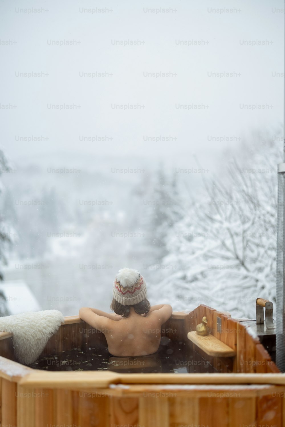 Donna che si rilassa nel bagno caldo all'aperto, sedendosi e godendo di una splendida vista sulle montagne innevate. Vacanze invernali in montagna, concetto di trattamento dell'acqua calda. Donna caucasica che indossa il cappello invernale