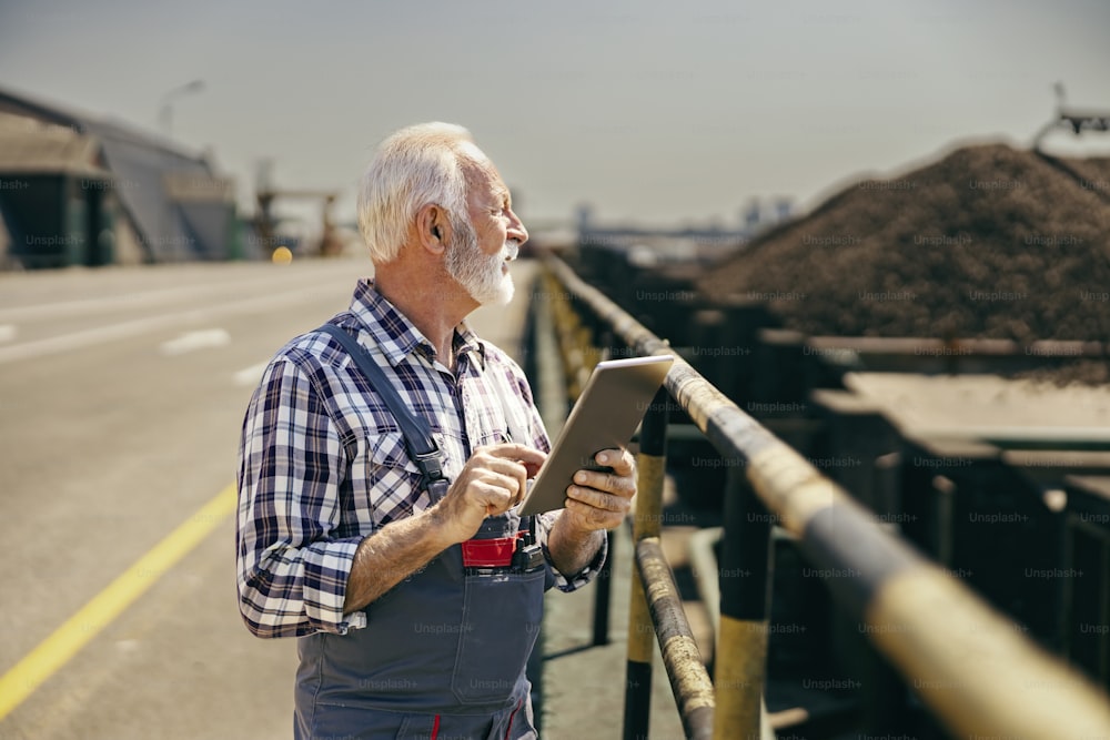 Control of works on construction site. A senior worker is standing on the highway with a tablet in his hands and checking on works.