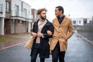 Stylish gay couple hug while walking together with eco shopping bag on some residential street on rainy weather. Caucasian and hispanic man wearing coats. Concept of homosexual lifestyle and fashion