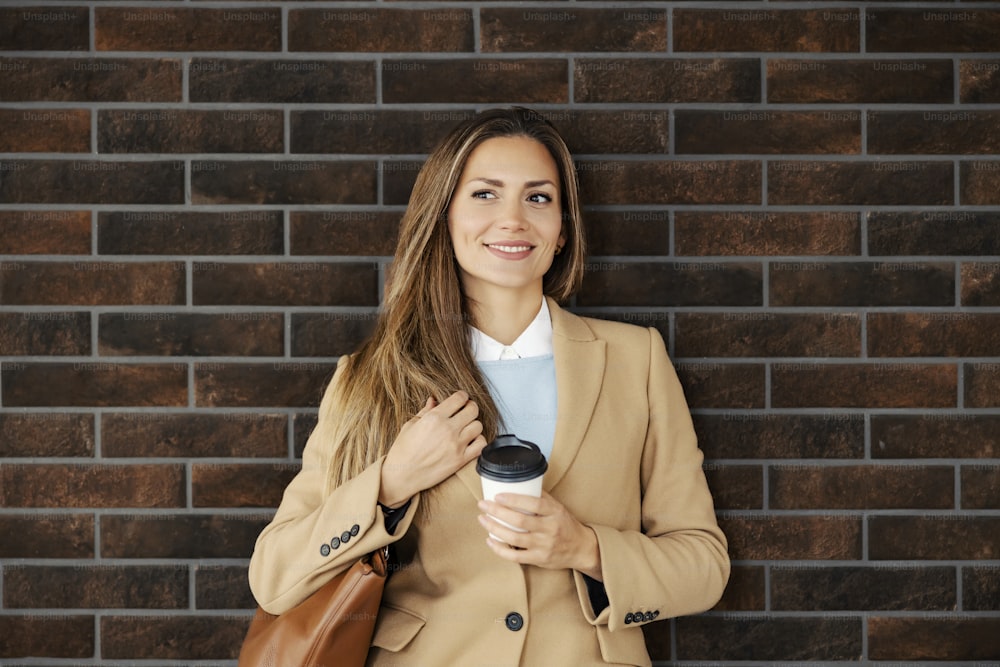 A woman in a coat standing on the street and holding takeaway coffee. A happy young woman in a coat standing against the brick wall and holding fresh coffee to go.