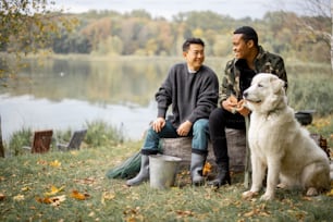 Multiracial male friends talking while sitting on fallen tree near Maremmano-Abruzzese Sheepdog on lake or river coast. Concept of rest and weekend in nature. Idea of friendship. Autumn day