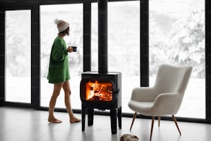겨울철 자연에 있는 현대 주택의 창문 근처 벽난로 옆에 컵을 들고 서 있는 여자. 집에서 겨울 분위기와 편안함의 개념. 산속의 오두막에서 레크리에이션에 대한 아이디어