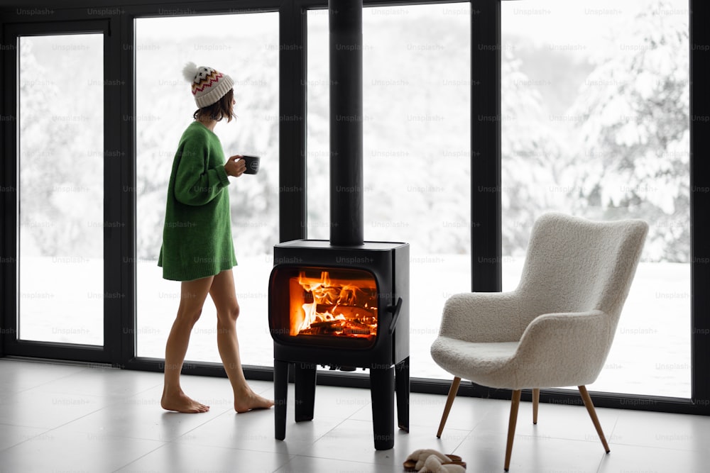 Woman standing with cup by the fireplace near the window at modern house on nature during winter time. Concept of winter mood and comfort at home. Idea of recreation in cabins on mountains