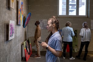 Vista lateral de una mujer joven con rastas que miran la pintura en la pared contra otros invitados de la galería de arte moderno