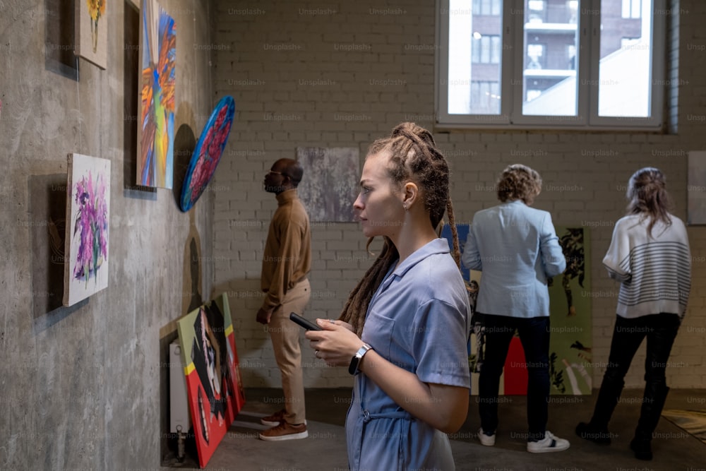 現代美術ギャラリーの他のゲストに対して壁に絵を見ているドレッドヘアの若い女性の側面図