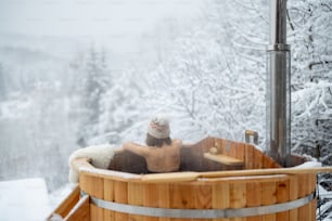 Femme se relaxant dans un bain chaud à l’extérieur, s’asseyant et profitant d’une belle vue sur les montagnes enneigées. Vacances d’hiver à la montagne, concept de traitement à l’eau chaude. Femme caucasienne portant un chapeau d’hiver