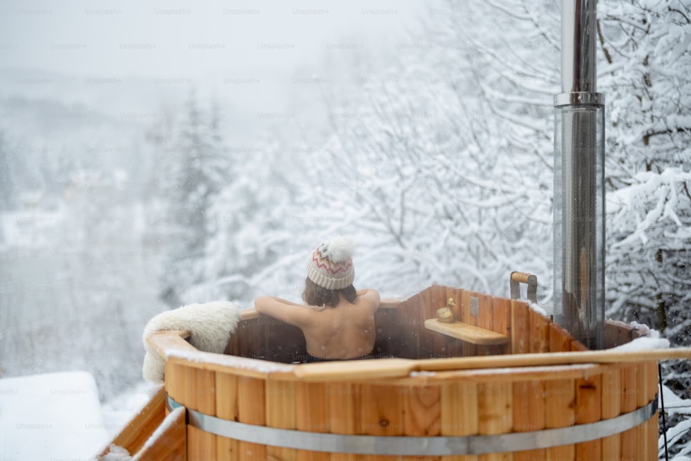 Frau entspannt sich im heißen Bad im Freien, lehnt sich zurück und genießt die schöne Aussicht auf die verschneiten Berge. Winterurlaub in den Bergen, Warmwasserbehandlungskonzept. Kaukasische Frau mit Wintermütze
