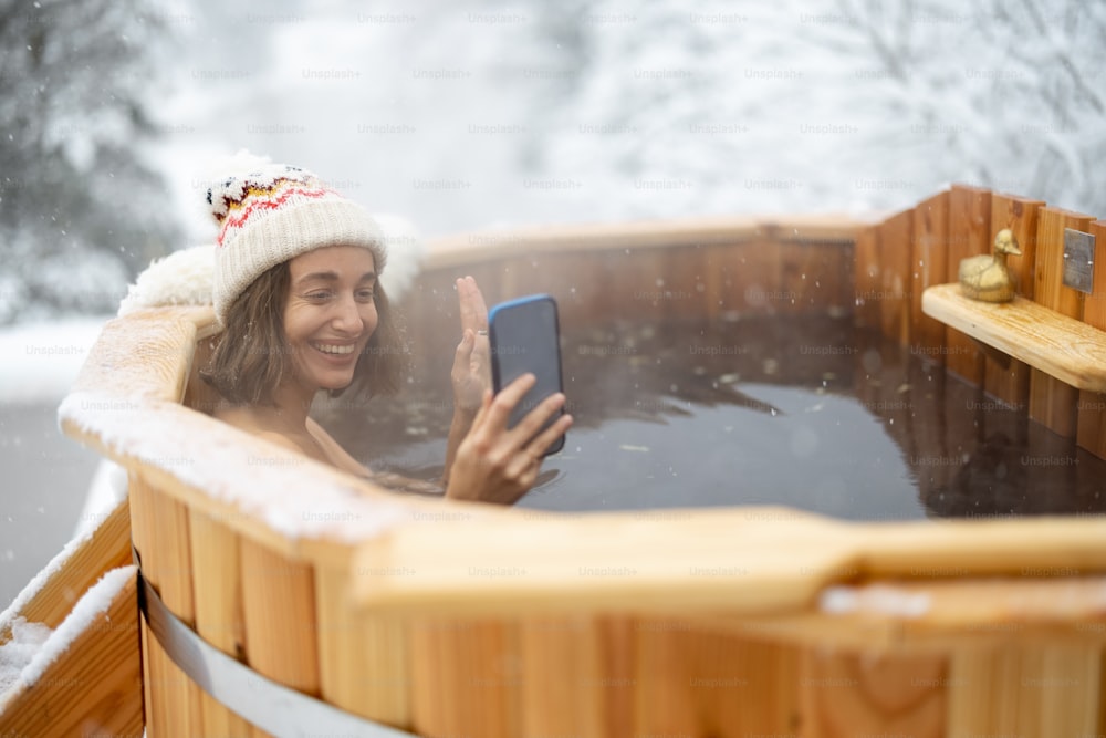 Happy woman relaxing in hot bath outdoors, talking on phone while sitting in thermal spa at snowy mountains. Winter recreation and water treatments concept. Idea of scandinavian lifestyle