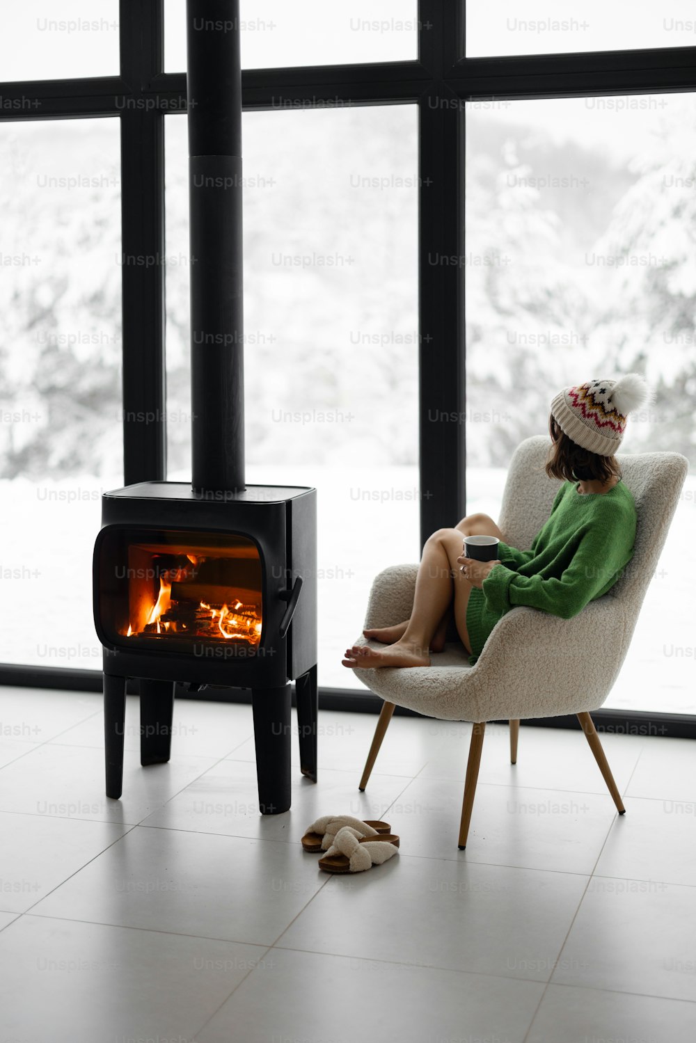 Woman sitting with cup on chair by the fireplace at modern house on nature during winter time. Concept of winter mood and comfort at home. Girl wearing hat and green sweater
