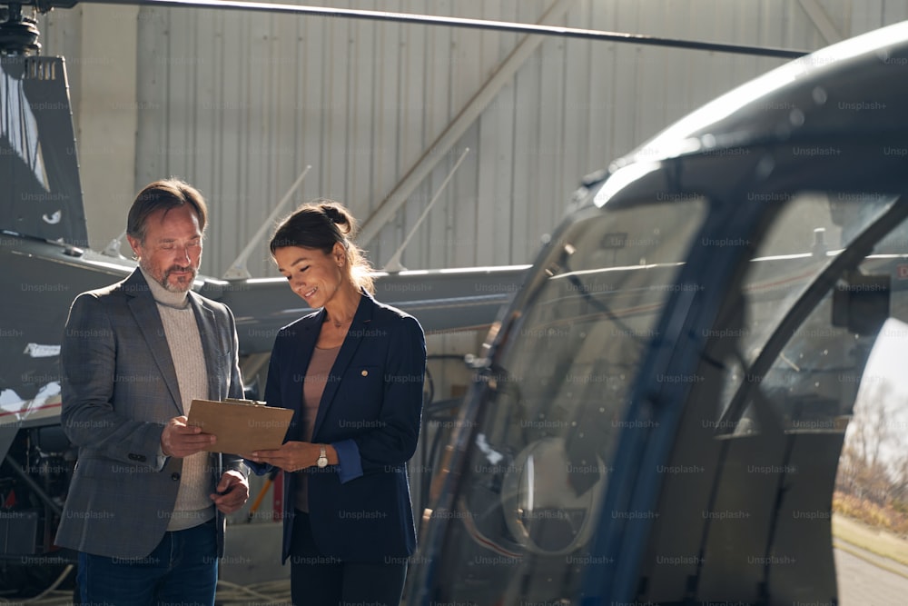 Une femme élégante souriante et un homme sérieux et concentré parcourant des documents parmi les hélicoptères sous hangar