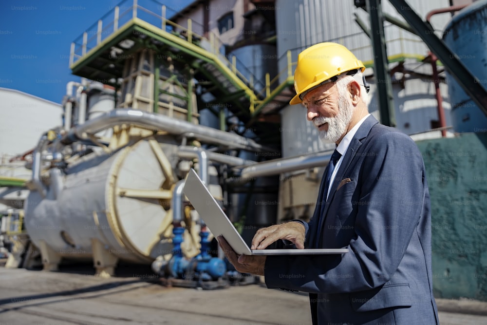 The oil industry and petroleum refinery. A successful senior refinery owner with a helmet on his head stands at a petroleum refinery and typing calculations on his laptop.
