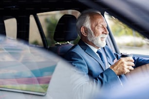 An old executive drinks coffee while driving a car. A happy senior businessman sits in his car and drives himself to work while finishing his morning takeaway coffee.
