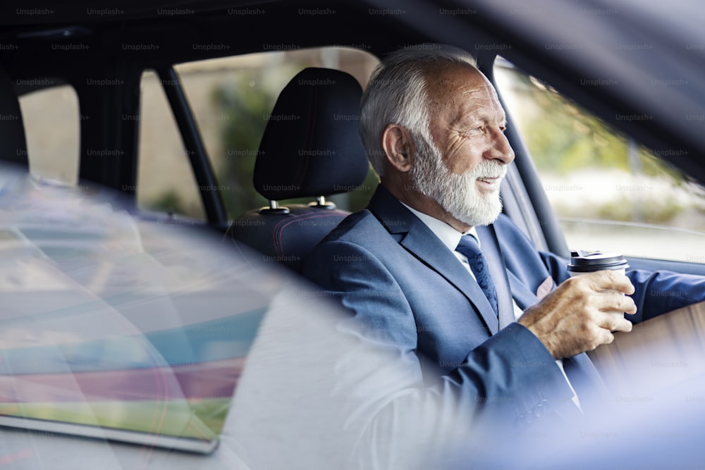 Un viejo ejecutivo toma café mientras conduce un coche. Un feliz hombre de negocios de la tercera edad se sienta en su automóvil y conduce al trabajo mientras termina su café matutino para llevar.