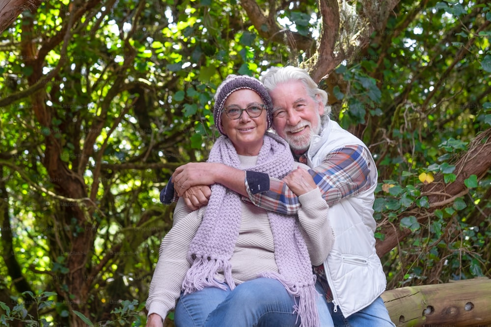 Happy senior couple enjoying mountain excursion in the woods embracing and smiling looking at camera - active retired people sitting in the shadow of trees