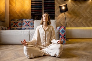 Relaxed woman feeling calm while meditating at modern apartment alone. Concept of mental health and home comfort. Caucasian woman wearing beige sleepwear