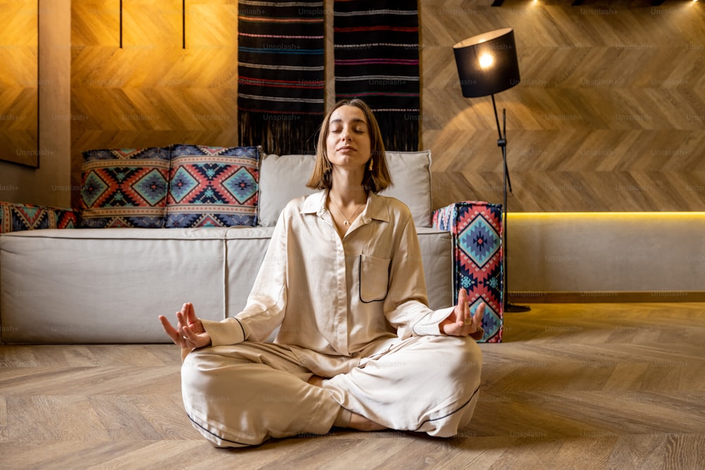 Relaxed woman feeling calm while meditating at modern apartment alone. Concept of mental health and home comfort. Caucasian woman wearing beige sleepwear