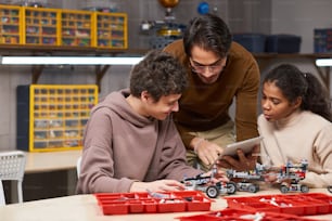 Giovane insegnante che aiuta i bambini a collegare i robot con la tavoletta digitale mentre sono seduti al tavolo