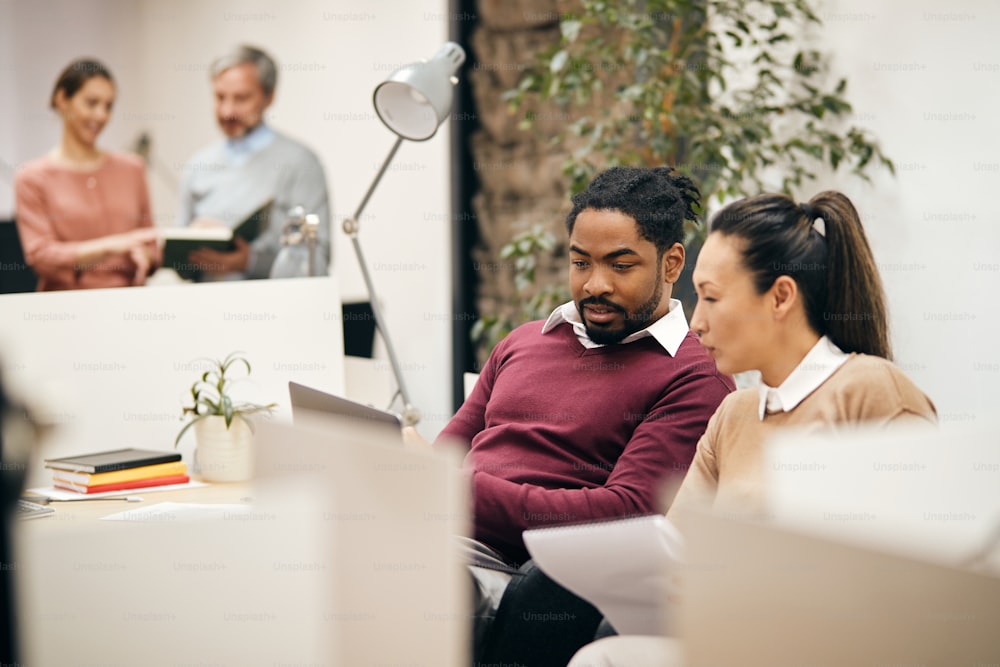 African American businessman reading an e-mail on laptop with female Asian coworker while working in the office.