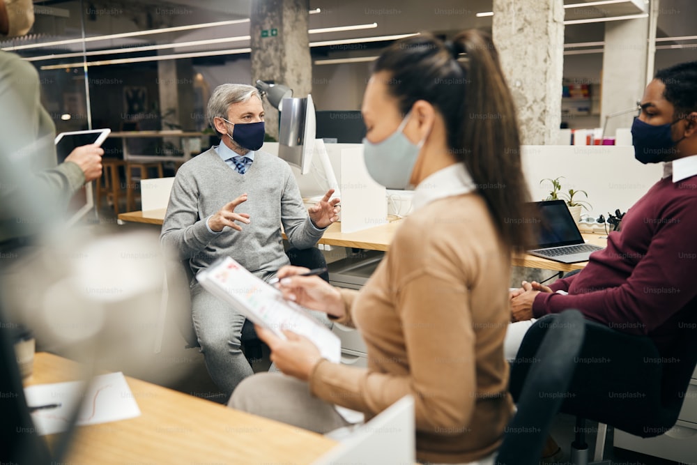 Männlicher CEO im Gespräch mit seinem Business-Team während eines Meetings im Büro. Sie tragen aufgrund der Coronavirus-Pandemie Schutzmasken.
