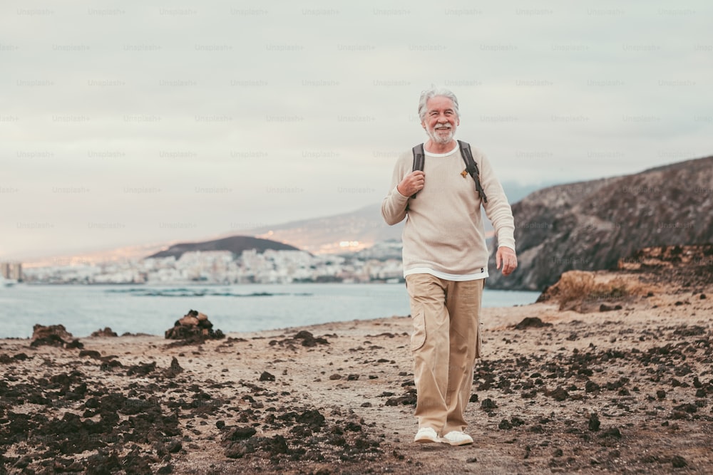 Old senior man walking on the cliff wearing backpack and smiling, enjoying sea excursion at sunset light. Active white-haired retired man