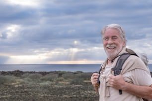Abuelo mayor feliz disfrutando de una excursión al aire libre en el mar sosteniendo una mochila. Horizonte sobre el agua y cielo dramático con rayos de sol