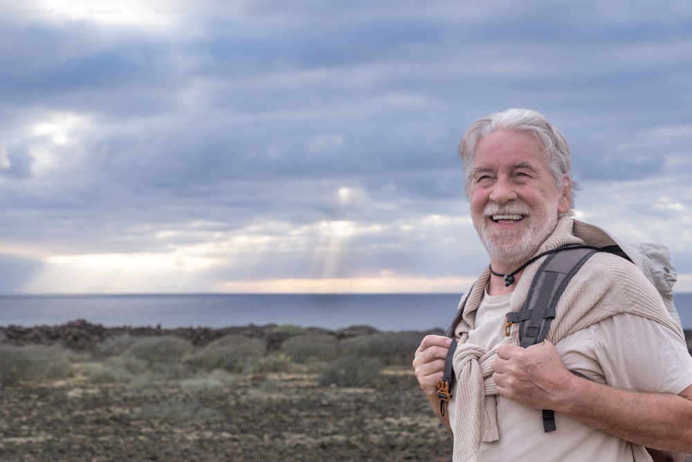 Avô sênior feliz desfrutando de excursão ao ar livre no mar segurando mochila. Horizonte sobre a água e céu dramático com raio de sol