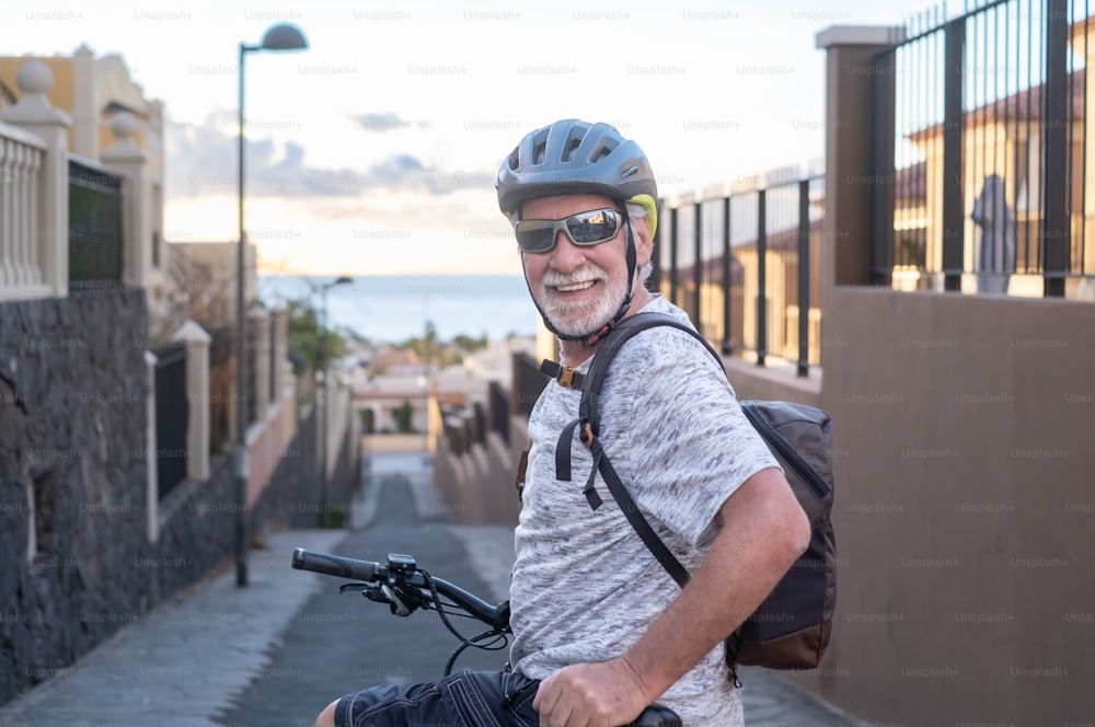 Active adult senior smiling man with sport helmet and sunglasses looking at camera cycling with his electric bicycle at sunset. Elderly man enjoying retirement and healthy lifestyle. Horizon over water in background
