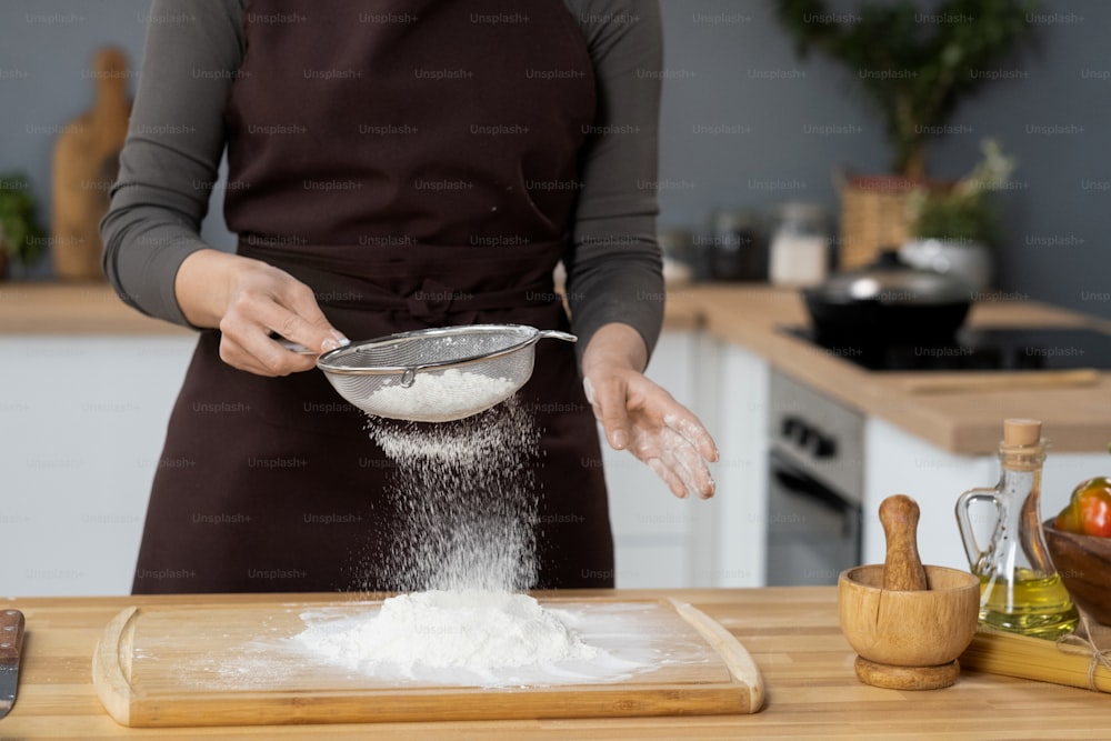 Donna contemporanea in grembiule che setaccia la farina dal tavolo della cucina di legno mentre prepara gli ingredienti per l'impasto e la pasticceria fatti in casa