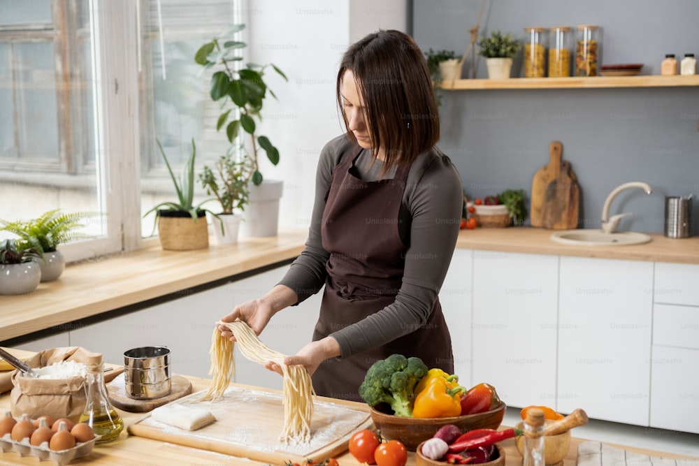 野菜とイタリアのパスタを準備しながら、台所のテーブルの上にゆでスパゲッティを保持している若いブルネットの女性