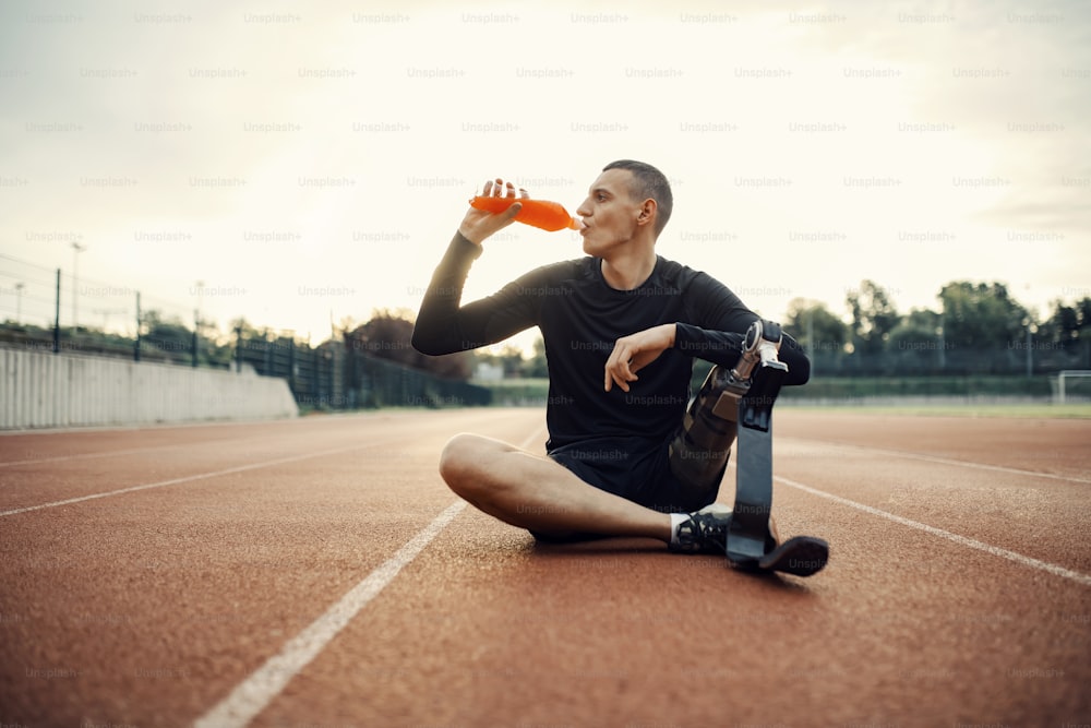 A thirsty runner with prosthetic leg sitting on running track at stadium and drinking refreshment.