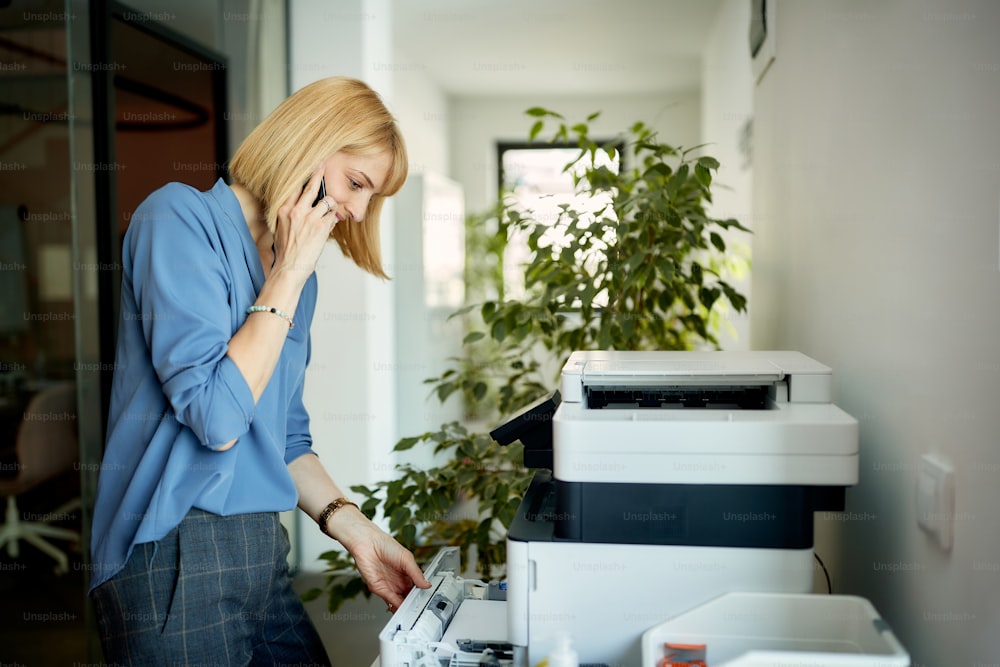 Donna d'affari sorridente che parla al telefono cellulare mentre usa la fotocopiatrice in ufficio.